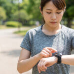 晴れた日に公園を走る日本人女性が、自身のスマートウォッチを見て心拍数を確認している様子。