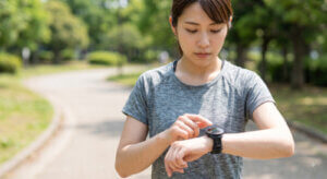 晴れた日に公園を走る日本人女性が、自身のスマートウォッチを見て心拍数を確認している様子。