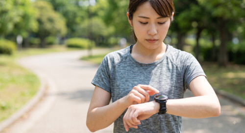 晴れた日に公園を走る日本人女性が、自身のスマートウォッチを見て心拍数を確認している様子。