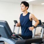 ジムの明るい有酸素エリアで、最新のランニングマシンを使い、真剣な表情でランニングをする日本人男性。