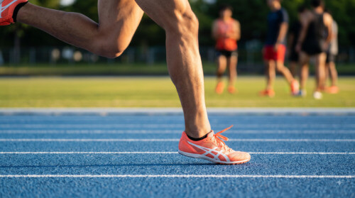 陸上競技場のブルーの新トラックで、オレンジと白の短距離用トレーニングシューズを履いた日本人男性スプリンターの右足が接地し、爆発的な反発力を生み出している、臨場感のあるクローズアップ写真。