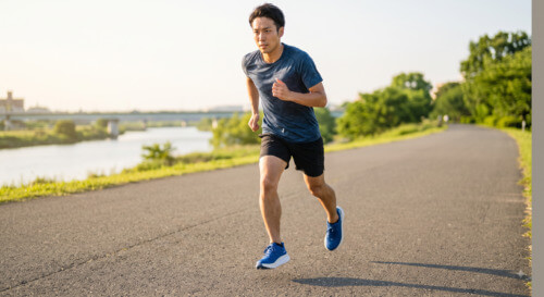 日夕の川沿いの舗装路を走る、厚底ランニングシューズを履いた日本人男性ランナーのダイナミックなストライド。