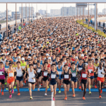 フルマラソンのスタートゲートを通過する日本人先頭ランナーと、背後に続く大勢の一般ランナーの海。上位4%のサブスリーの壁に挑む壮大な景色。
