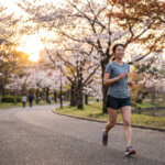 自然豊かな公園の小道を、笑顔でリラックスしてゆっくりとジョギングする日本人の女性ランナー。夕暮れ時の温かい光が差し込んでいる。