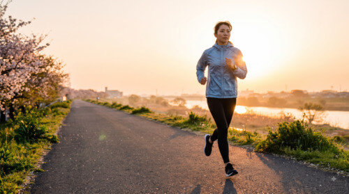 早朝の川沿いの小道を、朝日を浴びながら走る日本人の女性ランナー。健康的でモチベーションの高い様子。