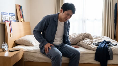 フルマラソン翌朝、激しい筋肉痛と疲労感に顔をしかめながら、ベッドの脇で太ももを押さえて立ち上がろうとする日本人男性ランナーの様子。